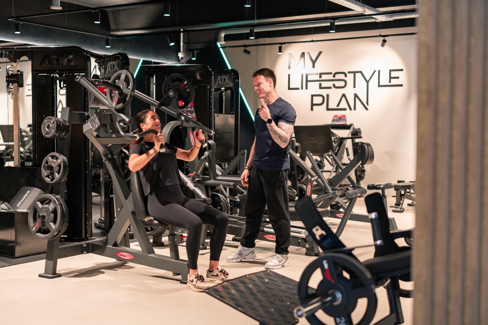 Een coach begeleidt een lid op een machine, met de MLP-wand erachter