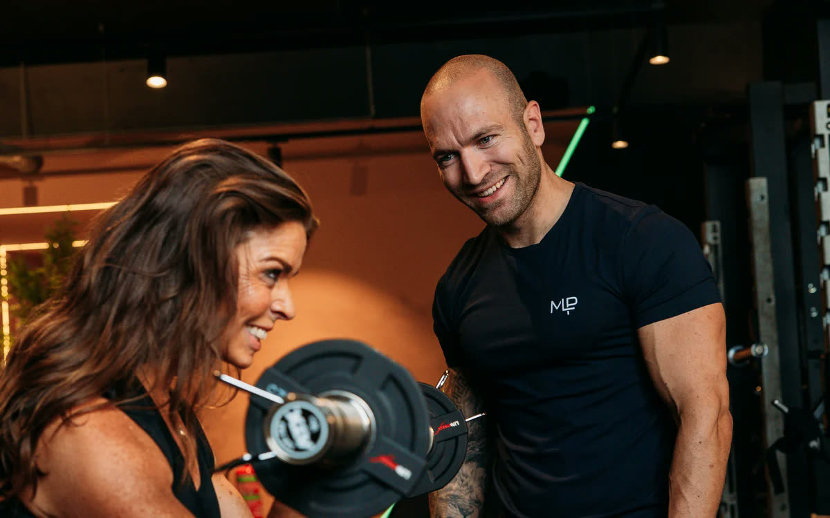 Een coach kijkt lachend mee terwijl een lid een curl met een halter doet