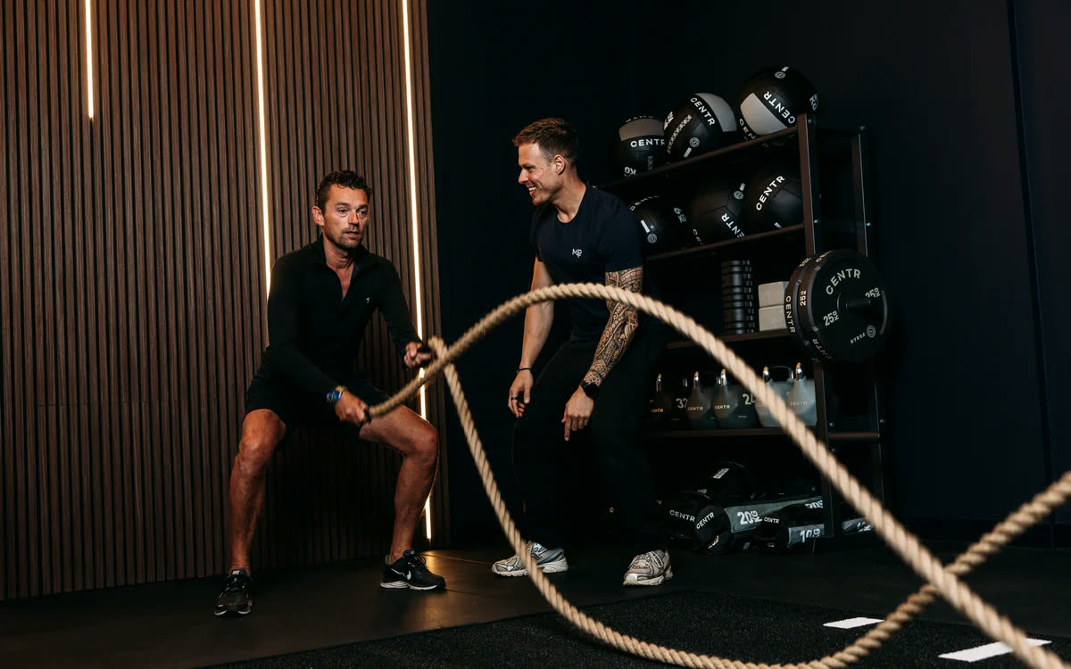 Een lid traint met een battle rope terwijl een coach meekijkt
