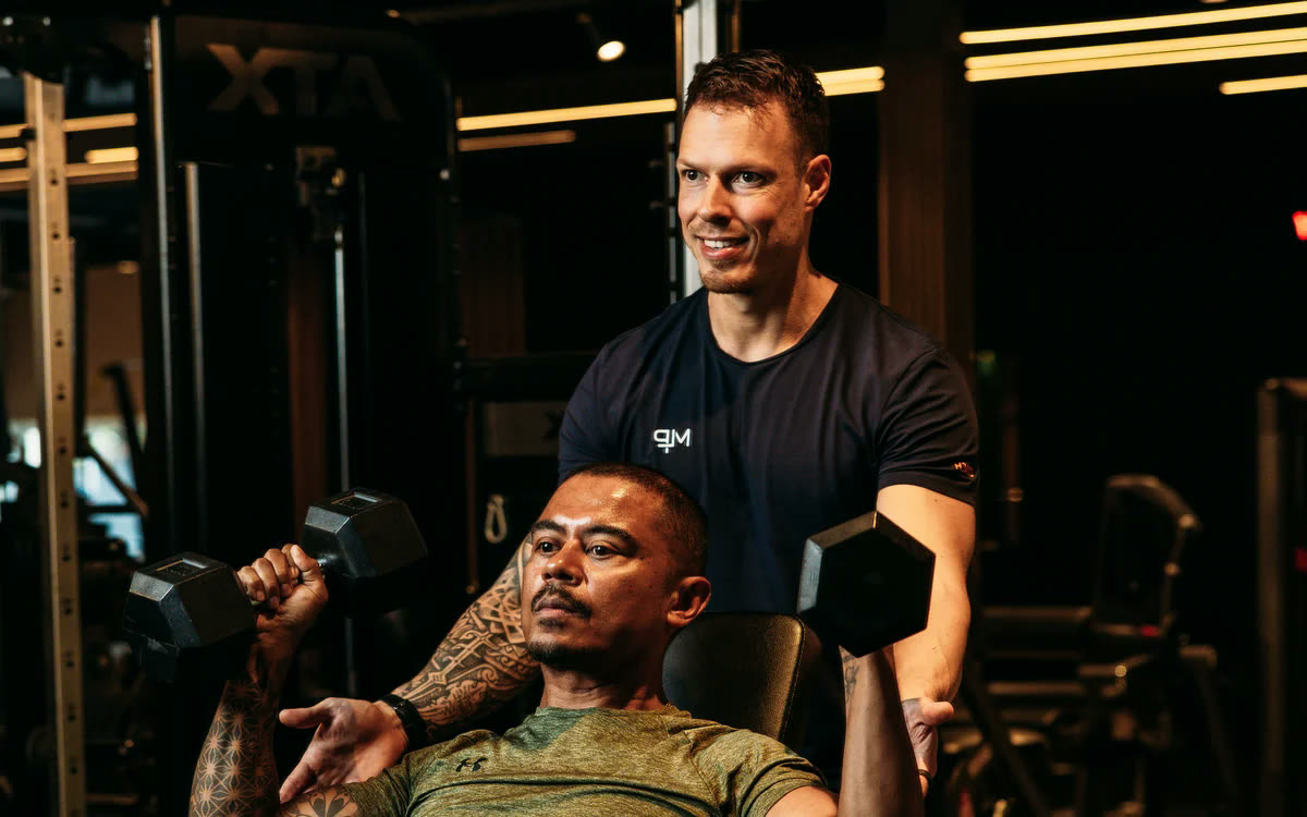 Een coach staat achter een lid dat dumbbells perst op een schuine bank